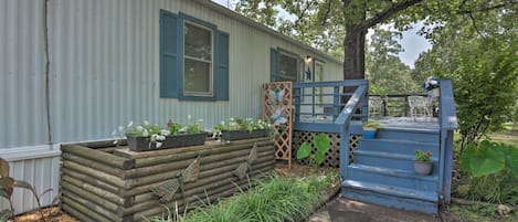 Una cabaña rústica con terraza de madera, un pequeño porche con barandal azul y un jardín con diversas plantas y flores.