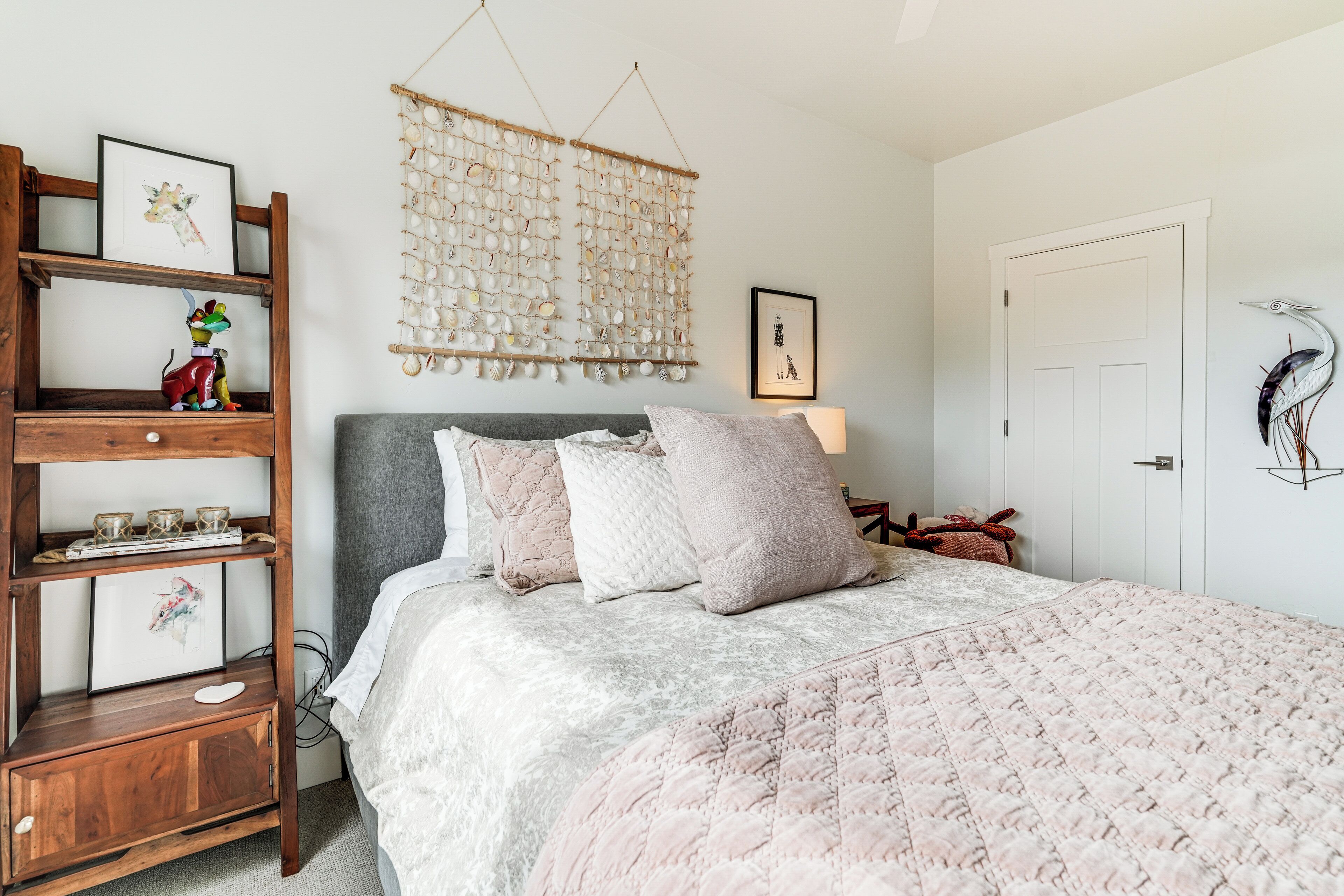 A bedroom with a bed, a wooden shelf with decorative items, a wall hanging, and a door.