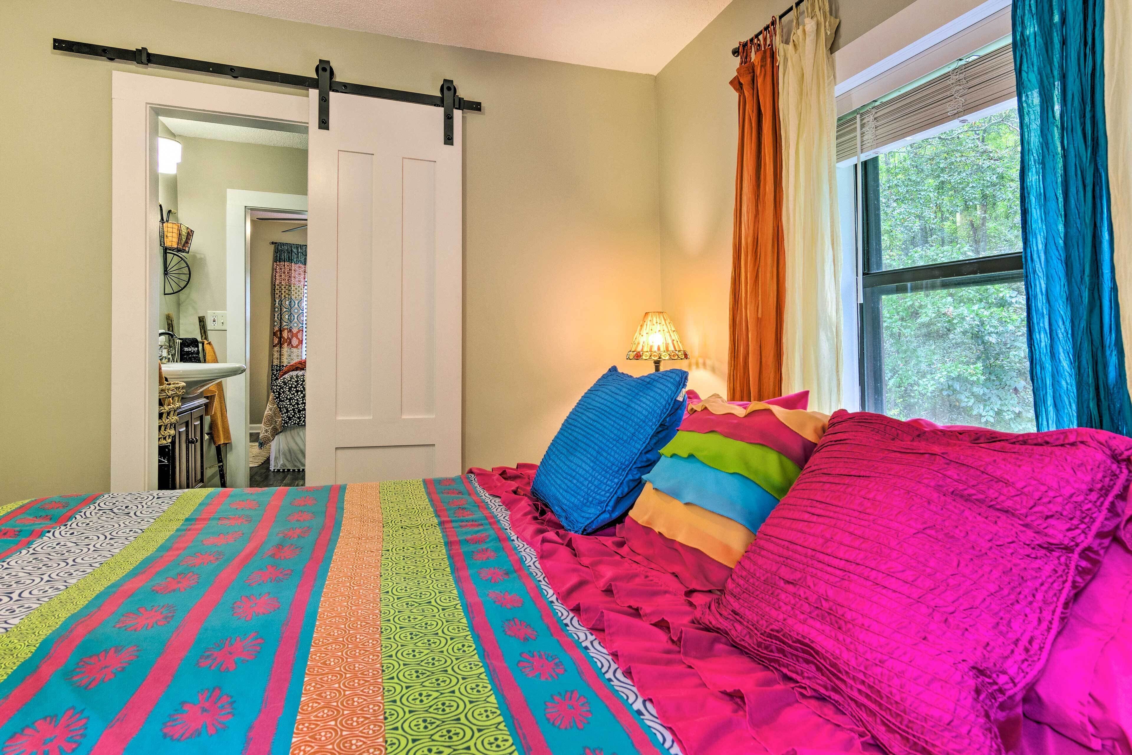 Un dormitorio con cama, ropa de cama colorida, una ventana con cortinas y una puerta que conduce a otra habitación.