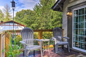 A wooden deck with two chairs and a small table, surrounded by greenery and a house in the background.