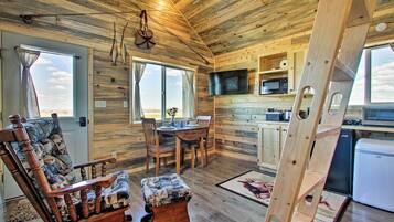 A wooden cabin interior with a dining area, a TV mounted on the wall, and a kitchen area with a microwave and a refrigerator.