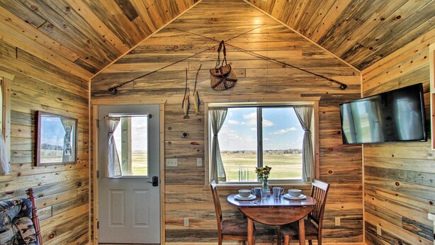 A wooden cabin interior with a dining area, a TV mounted on the wall, and a door leading outside. - Pano Views! Custom Cabin on Rocky Mountain Front (Augusta)