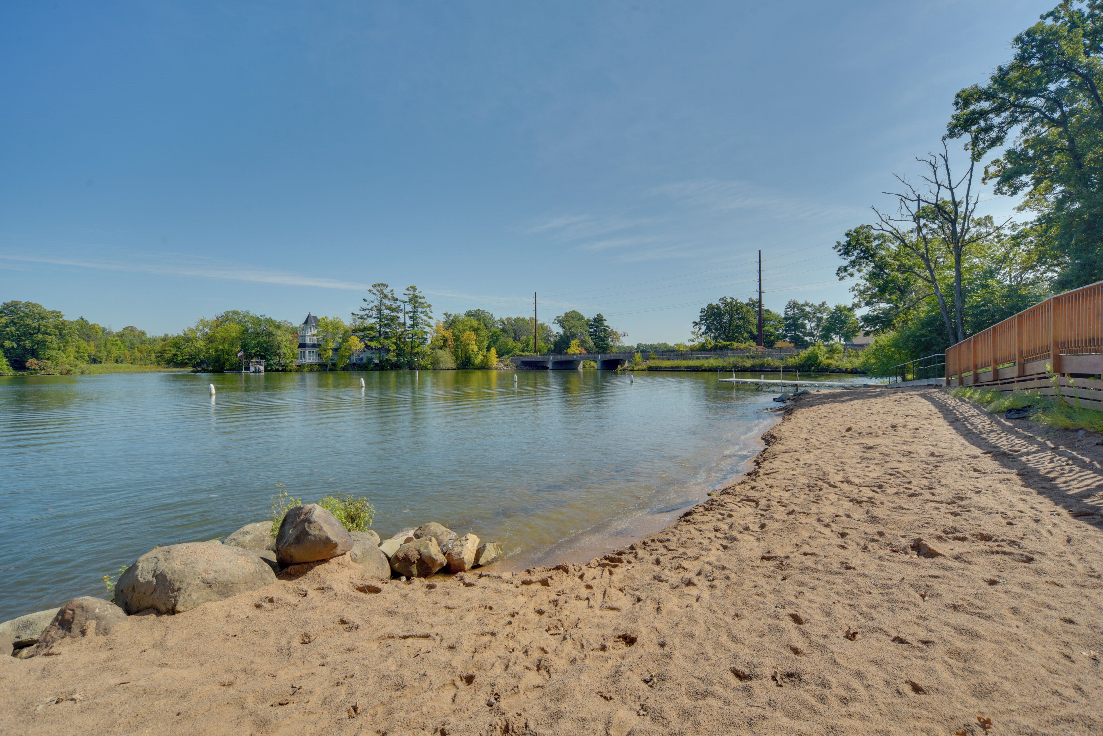 A sandy beach with a calm lake, a wooden bridge, and trees in the background.