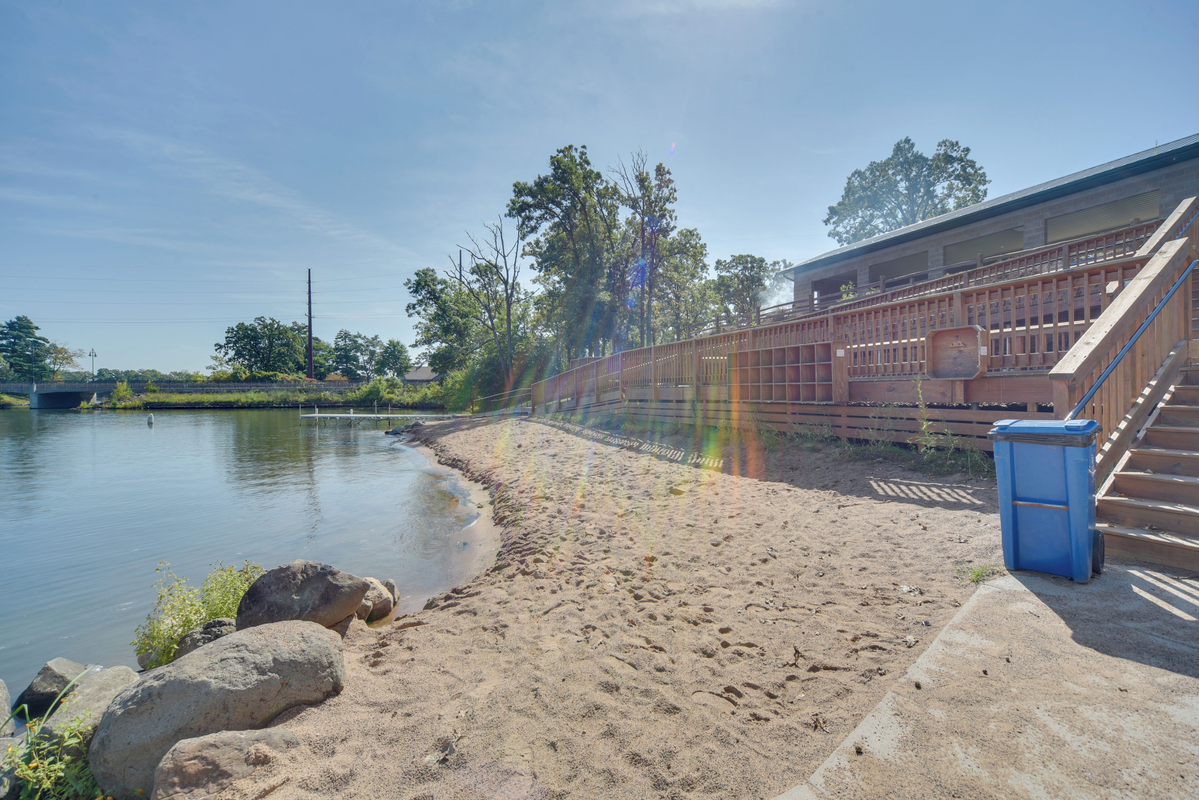 A riverside walkway with a wooden fence, a blue trash bin, and a building with a sloped roof.