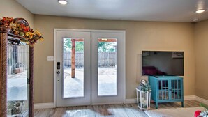 A room with a double door leading to an outdoor area, a television mounted on the wall, and a small blue cabinet. - Walk to Texas Tech! Cozy Lubbock Gem w/ Patio (Lubbock)