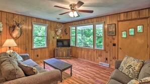 A room with wooden paneled walls, a ceiling fan, a television, a fireplace, and large windows with a view of trees.