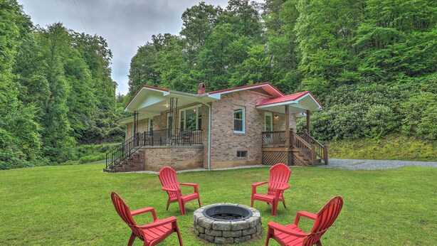 A house with a stone exterior, a covered porch, and a fire pit with red chairs in the yard.