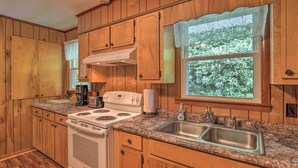 A kitchen with wooden cabinets, a granite countertop, a double sink, a stove, and a window with a view of greenery.