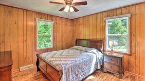 A bedroom with wooden paneling, a bed with a patterned bedspread, a nightstand with a lamp, and a ceiling fan.
