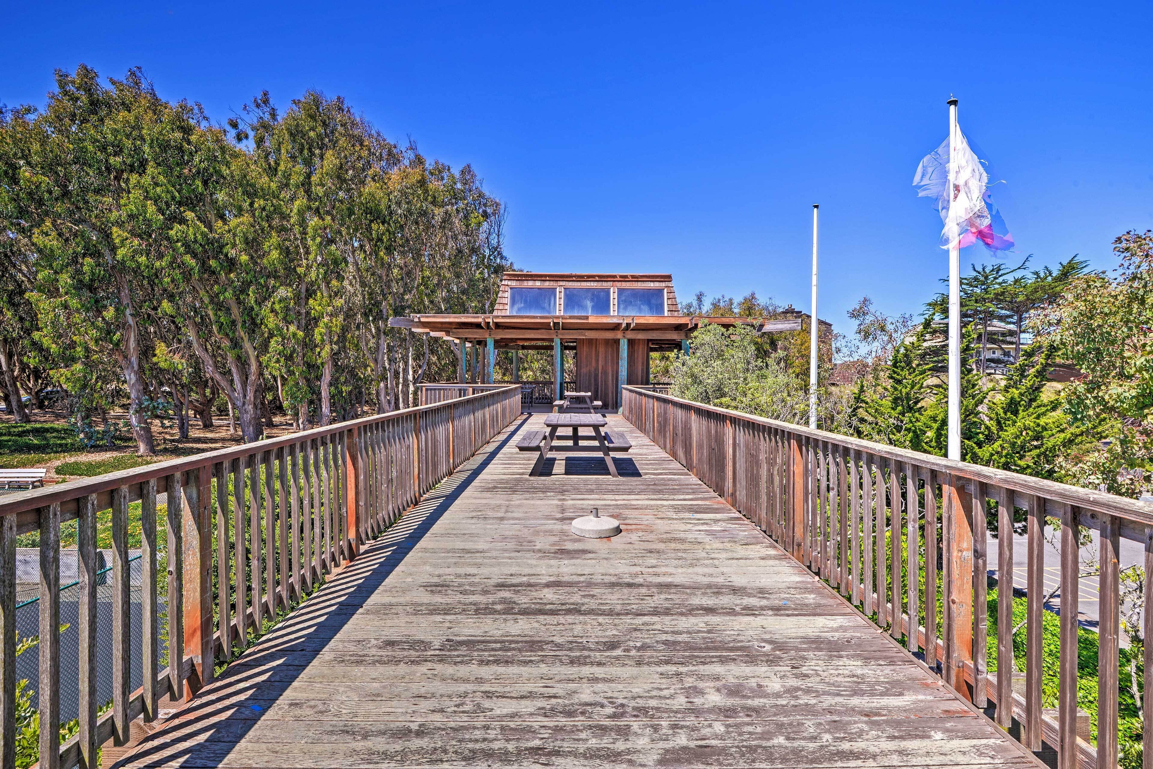 A wooden pedestrian bridge with a picnic table in the center, leading to a building with a flat roof and large windows.