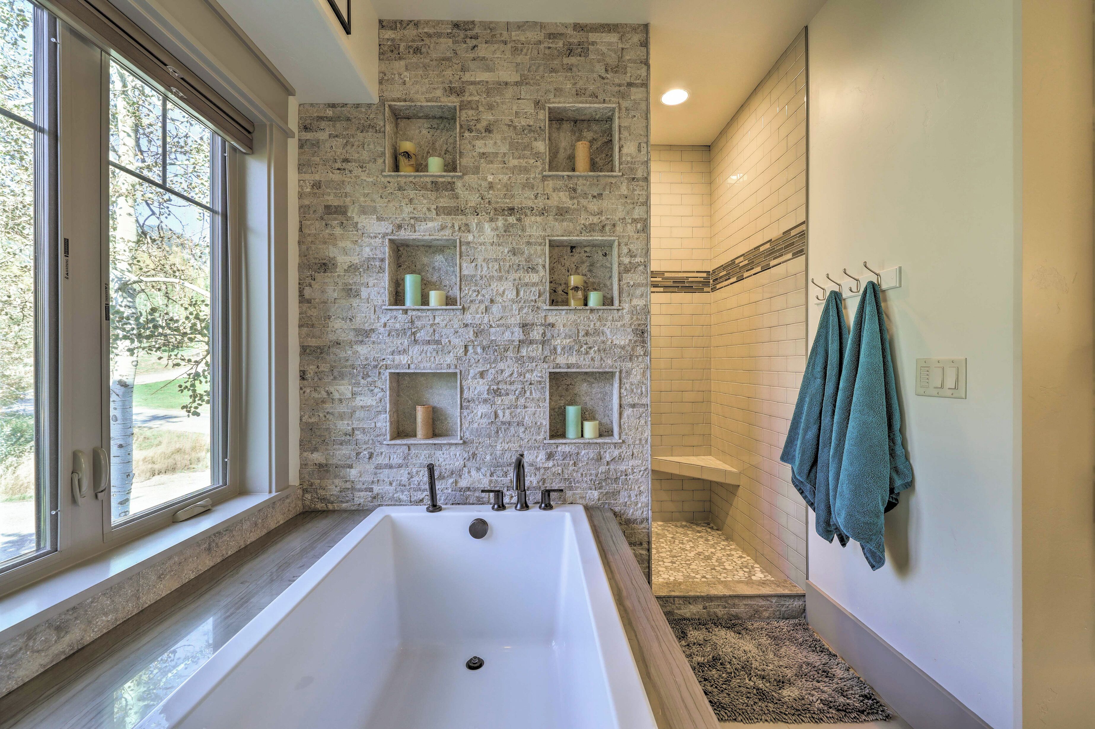 A bathroom with a large bathtub, a window with blinds, a brick wall with shelves, and a towel rack.