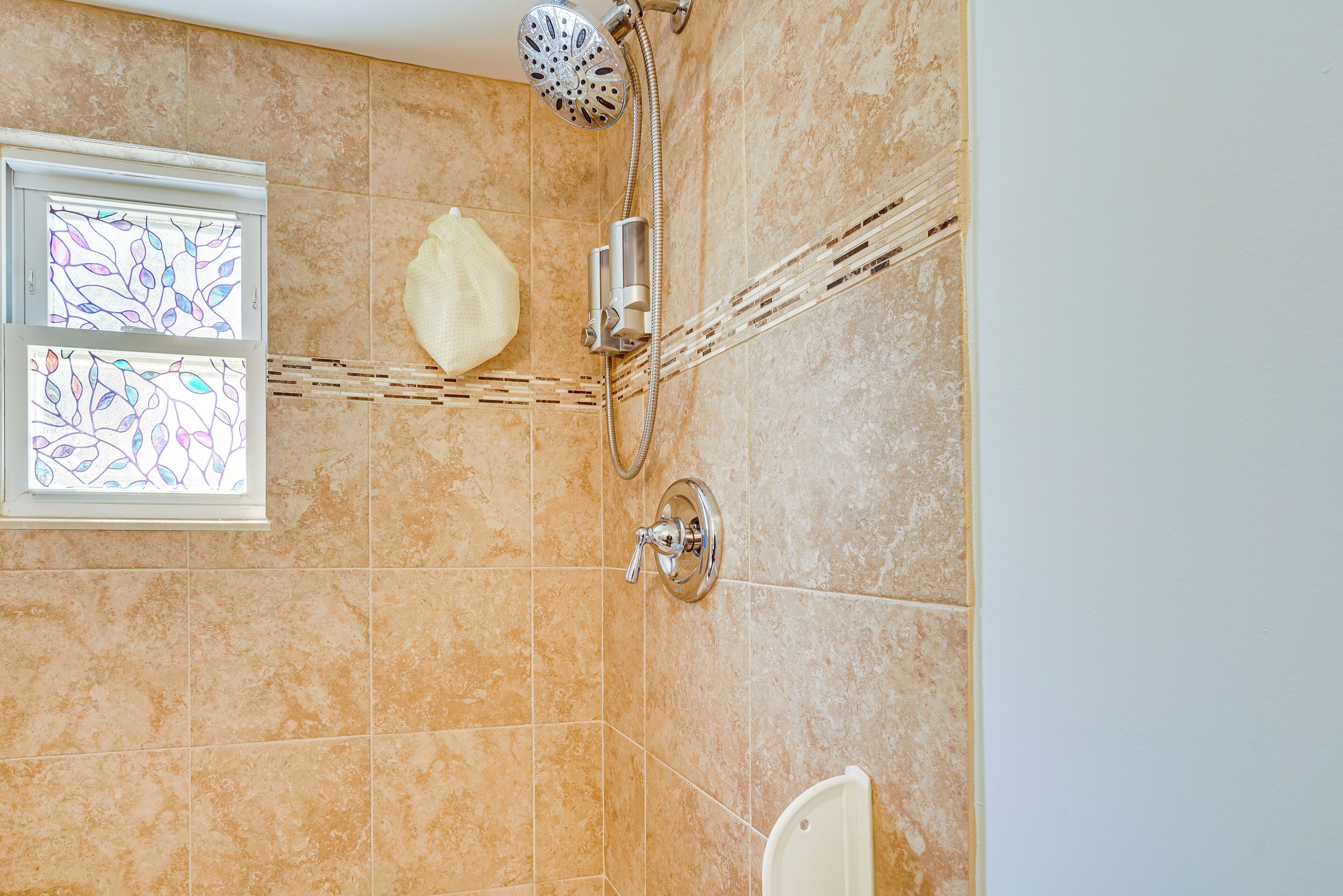 A bathroom with beige tiles, a shower with a handheld showerhead, and a window with stained glass.