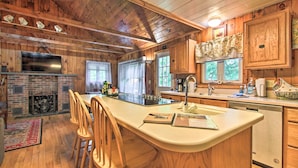A wooden cabin interior with a fireplace, wooden dining table with chairs, a kitchen area with a sink and counter, and a large window. - Ludlow Home w/ Hot Tub, Near Okemo Resort! (Ludlow)