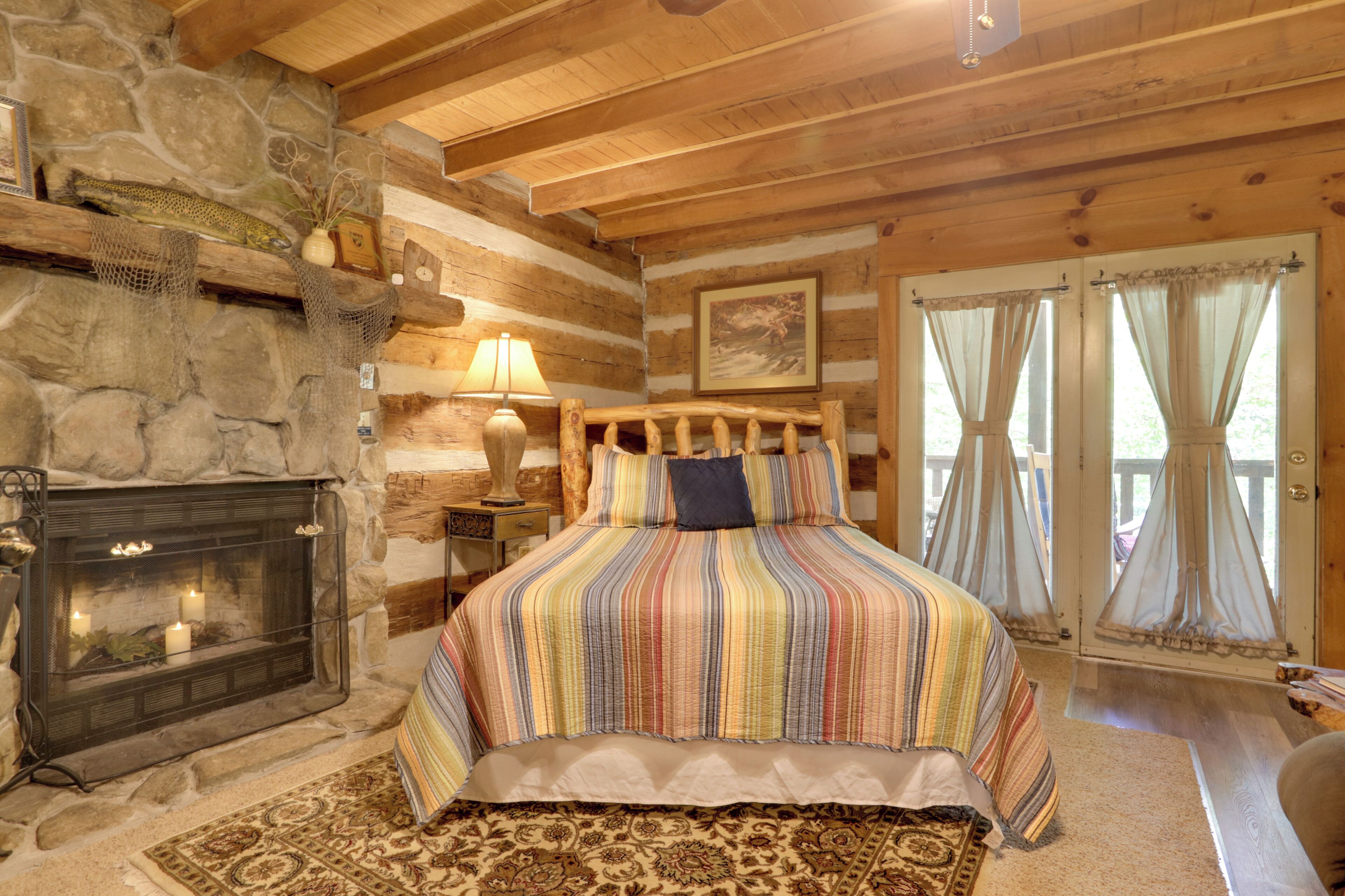 A cozy log cabin bedroom with a stone fireplace, a bed with a striped bedspread, and a view through a glass door.