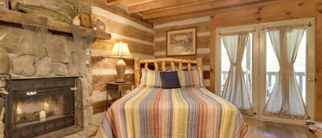 Une chambre confortable dans un chalet en rondins, avec une cheminée en pierre, un lit recouvert d’une courtepointe à rayures et une vue à travers une porte vitrée.