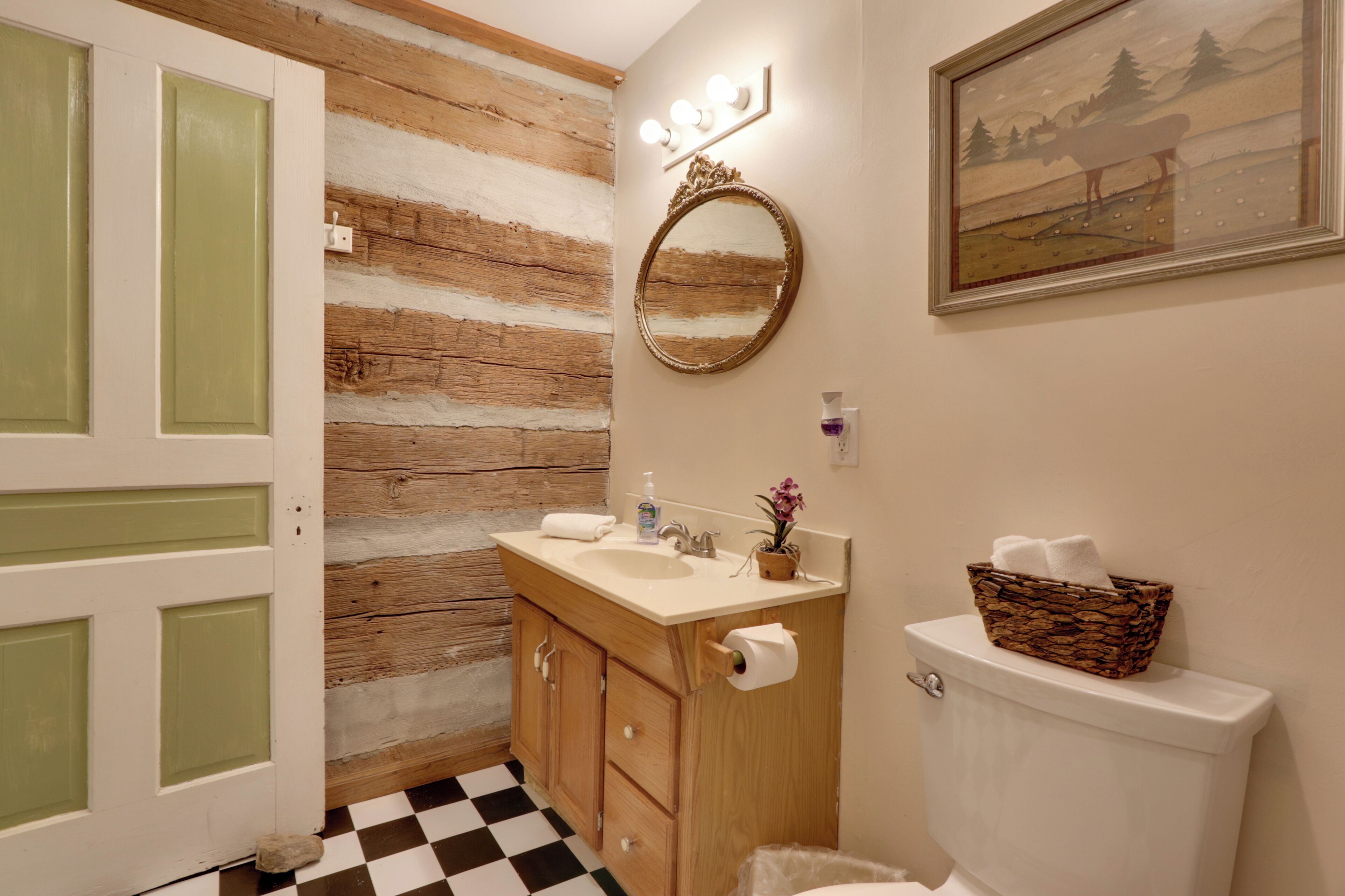 A bathroom with a wooden wall, a sink with a mirror, and a painting above the toilet.