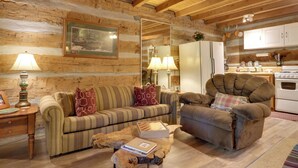 A cozy log cabin interior with a wooden-framed ceiling, a striped sofa, a plush armchair, a wooden coffee table, and a kitchen area with white cabinets and a stove. - 3 Mi to Natl Park: Townsend Apt on Little River! (Townsend)