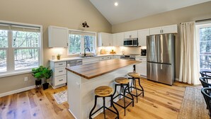 A modern kitchen with white cabinets, a stainless steel refrigerator, a wooden island with stools, and large windows offering a view of trees. - Chic Wisconsin Retreat w/ Deck, Grill & Fire Pit! (Wisconsin Dells)