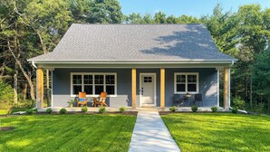 A single-story house with a grey roof and white siding, a front porch with two chairs, and a concrete pathway leading to the entrance. - Chic Wisconsin Retreat w/ Deck, Grill & Fire Pit! (Wisconsin Dells)