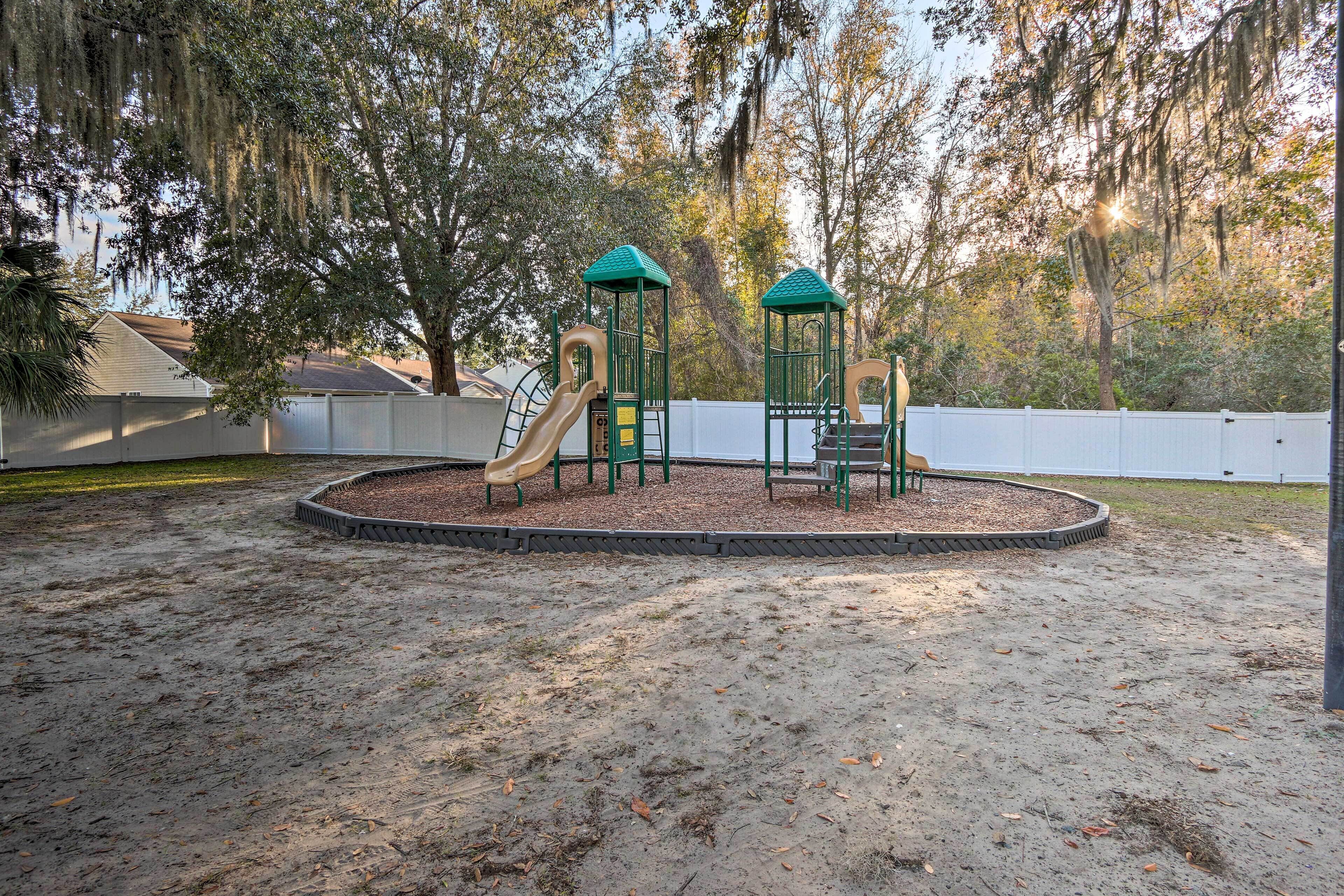 A playground with a slide and climbing structures, surrounded by a sandy area and trees.