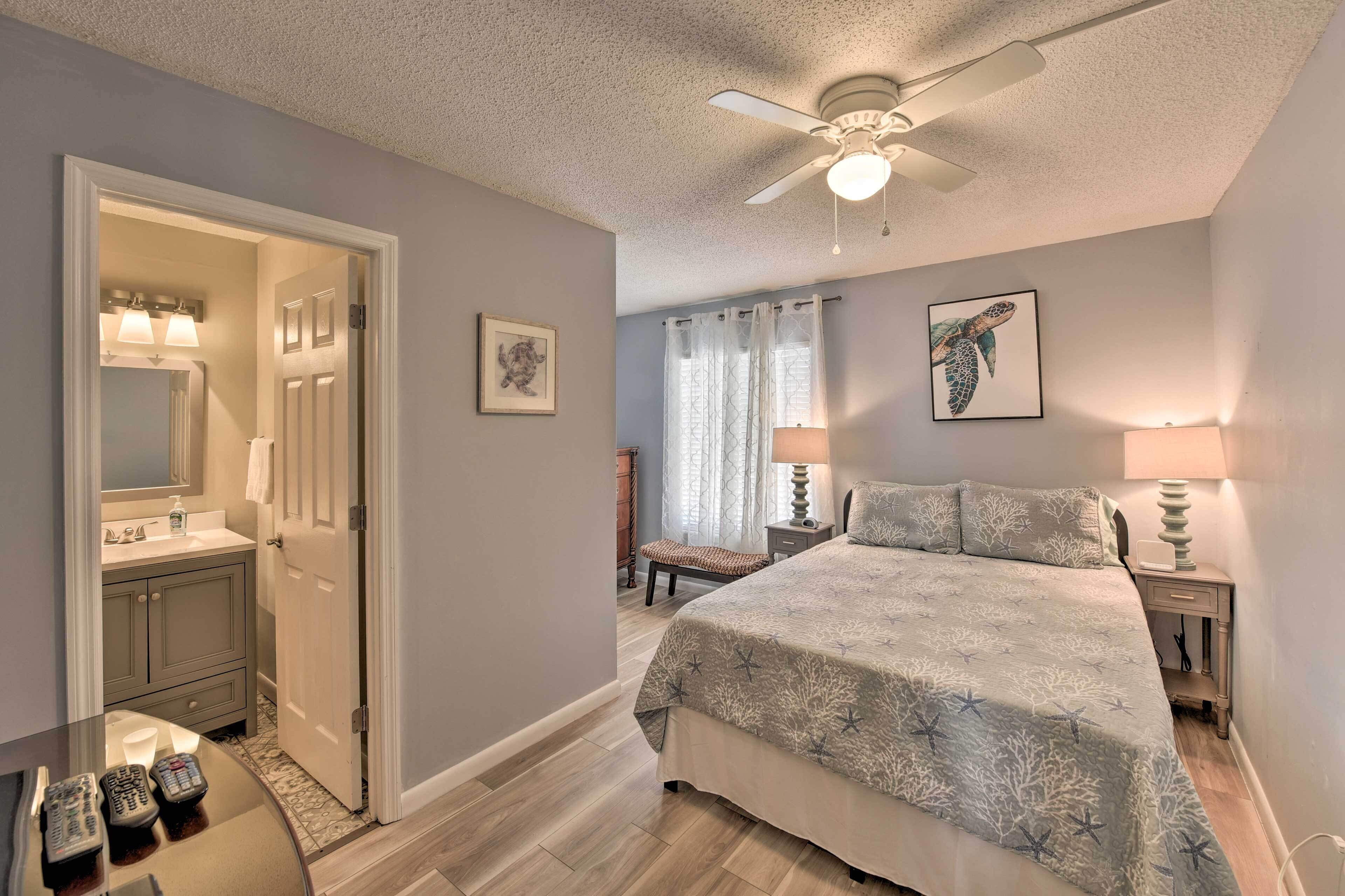 A bedroom with a bed, bedside tables, a nightstand, a ceiling fan, and a bathroom with a vanity, mirror, and towel rack.