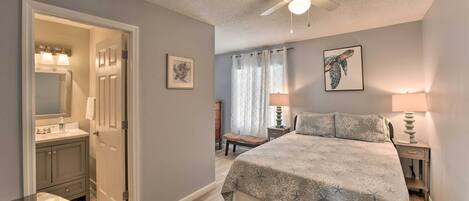 A bedroom with a bed, bedside tables, a nightstand, a ceiling fan, and a bathroom with a vanity, mirror, and towel rack.