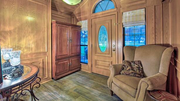 Una habitación con paneles de madera, un gran ventanal arqueado, una mesa redonda con una lámpara y un sillón con cojines.