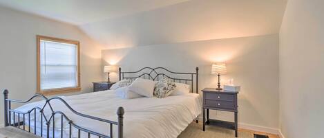 A bedroom with a metal bed frame, white bedding, two bedside tables with lamps, a wooden bench, and a window with blinds.
