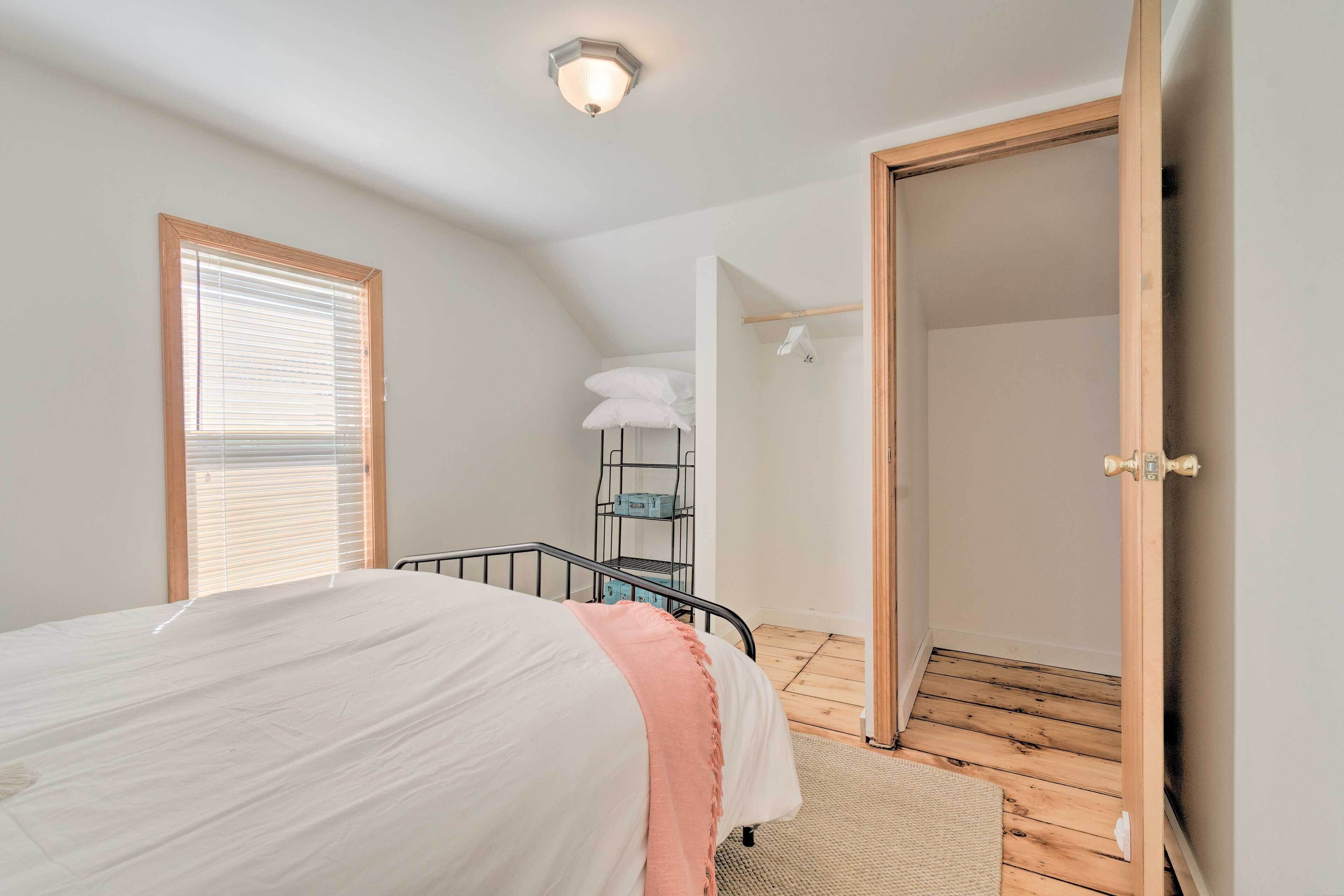 A bedroom with a bed, a wooden floor, a window with blinds, a wardrobe, and a door leading to another room.