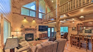 A spacious log cabin interior with a fireplace, a large sofa, a dining area, and a loft with a wooden staircase. - Island Park Cabin w/ Stunning Forest Views! (Island Park)