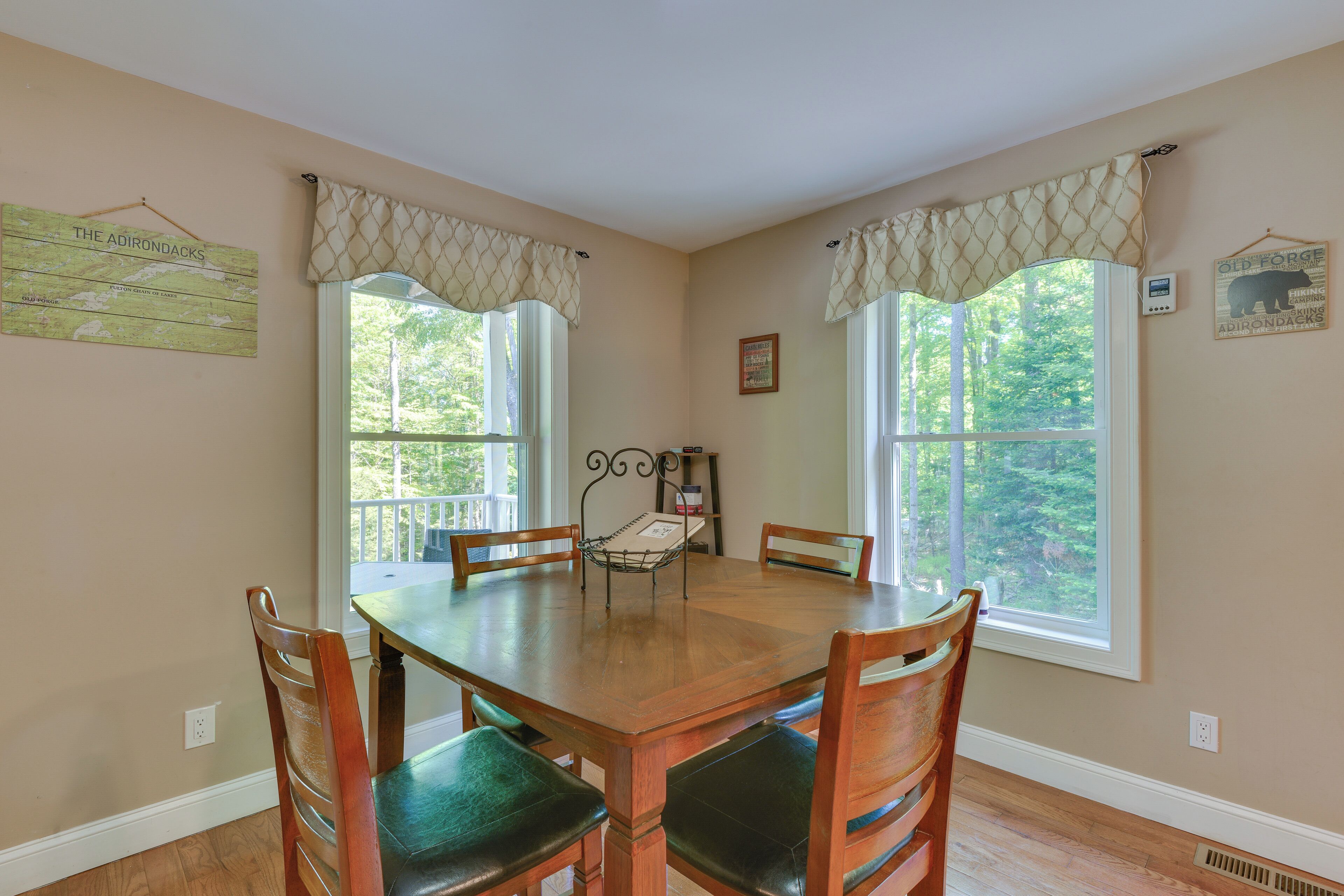 A dining room with a wooden table and chairs, large windows with curtains, a map on the wall, and a thermostat.