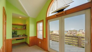 Una habitación con un gran ventanal corredero que da a una terraza de madera con vistas a la playa y edificios. Se observa una estantería empotrada con libros y un jarrón con flores.