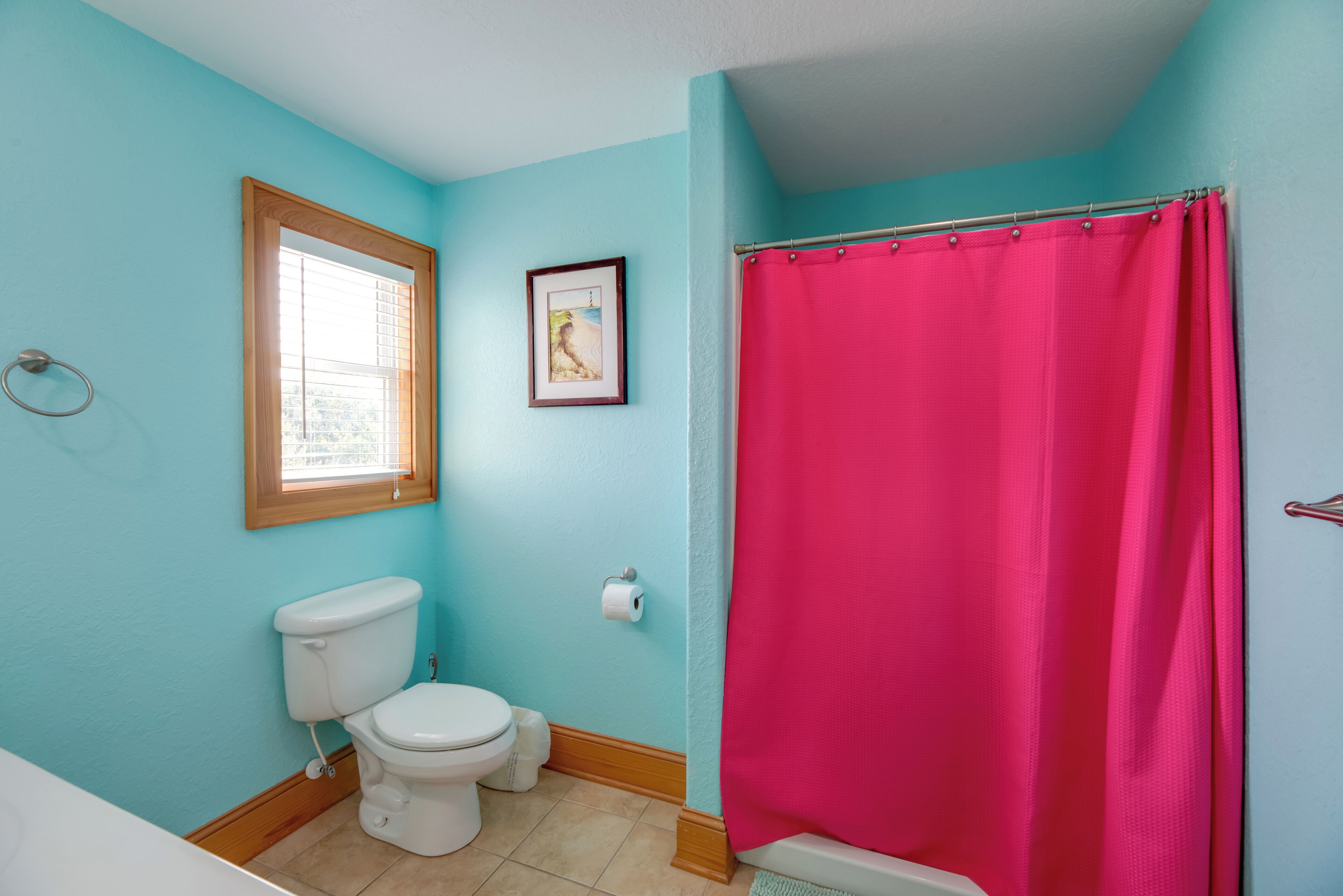 A bathroom with a toilet, a window, a shower curtain, and a framed picture on the wall.