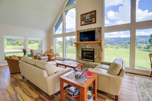 A spacious living room with a stone fireplace, large windows offering a view of a green landscape, and a high ceiling.