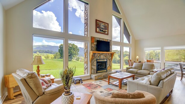 Amplia sala de estar con chimenea de piedra, ventanas grandes y vistas a un paisaje verde.