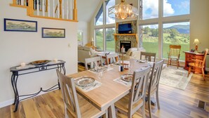 Un comedor con mesa de madera dispuesta para seis personas, chimenea y ventanas amplias con vistas al exterior.