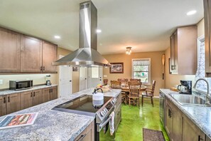 A kitchen with a central island, stainless steel appliances, wooden cabinets, and a dining area with a table and chairs. - Brand New Cotter Home, Walk to Trout Fishing! (Cotter)
