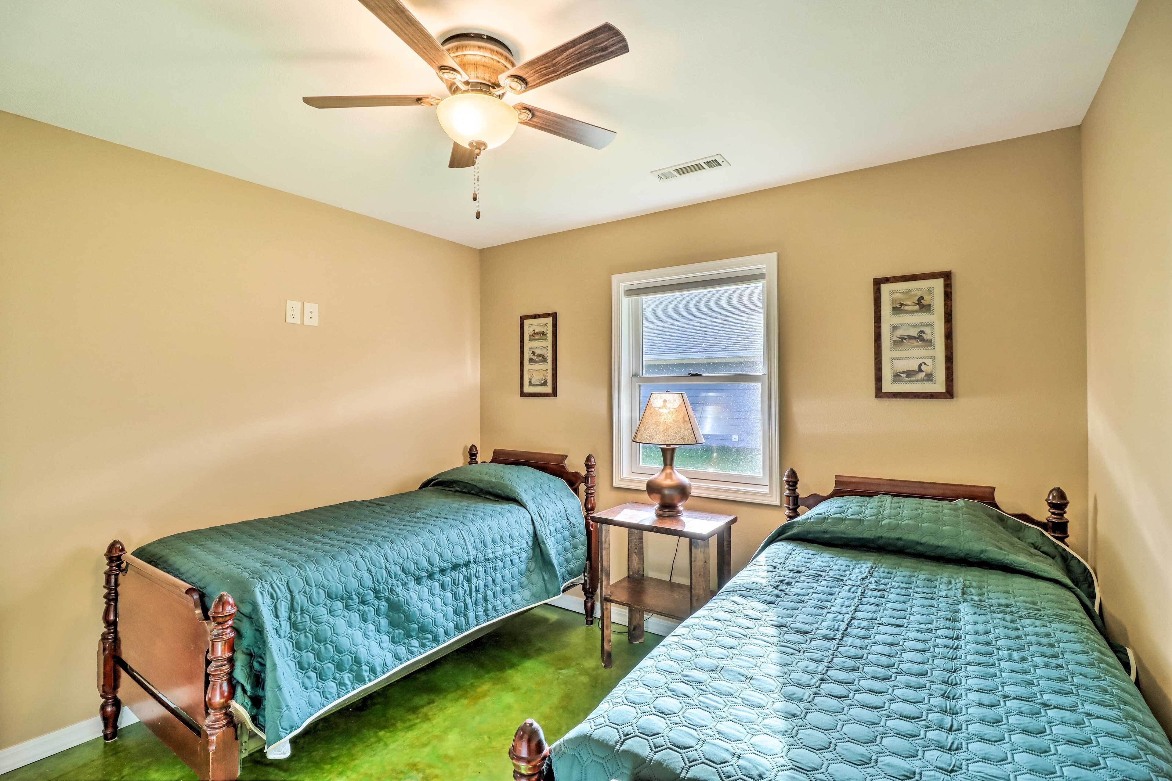 A bedroom with two wooden beds, a ceiling fan, a window with blinds, and a small table with a lamp.