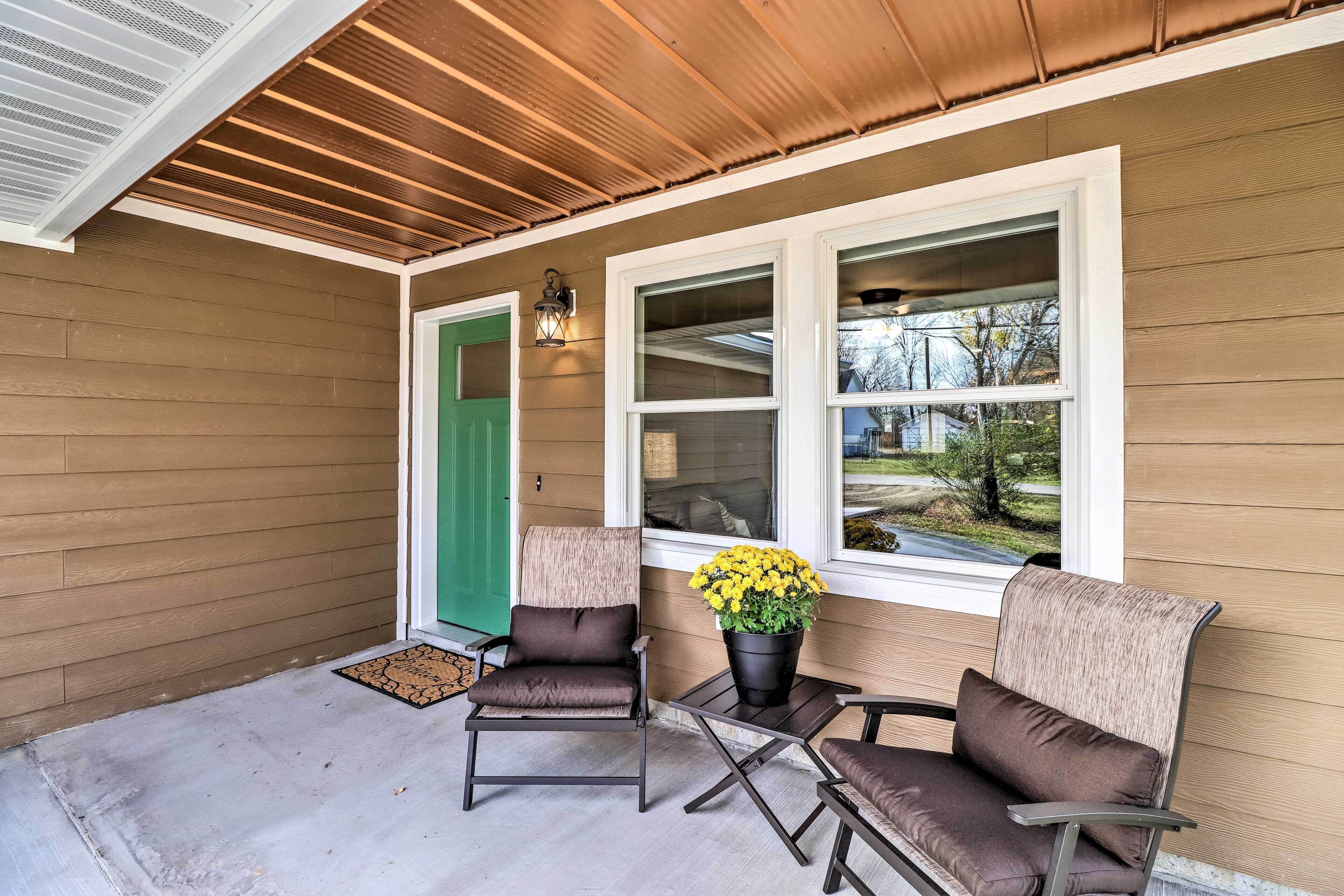 A covered porch with two chairs and a table, a green door, and a window with a view of the outside.