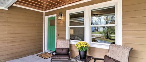 A covered porch with two chairs and a table, a green door, and a window with a view of the outside.