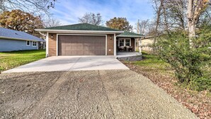 A single-story house with a brown garage door, a covered porch, and a gravel driveway. - Brand New Cotter Home, Walk to Trout Fishing! (Cotter)