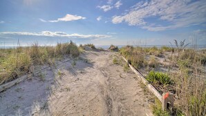 Een zandpad dat naar een strand leidt, met weinig beplanting en een houten hek aan één kant.