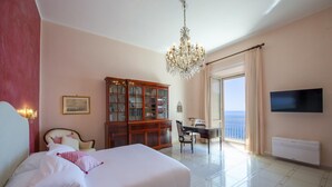 Un dormitorio con una cama grande, un escritorio con silla, una estantería, una araña y un televisor, además de vistas al mar.