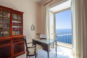 A room with a glass table, a wooden desk, a chair, a bookshelf, and a view of the ocean through a large window. - Villa Arte- Luxurious Villa in Ravello (Minori)