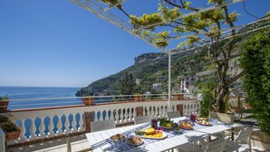 Una terraza con una mesa dispuesta para una comida, con vistas a la costa y una montaña al fondo.