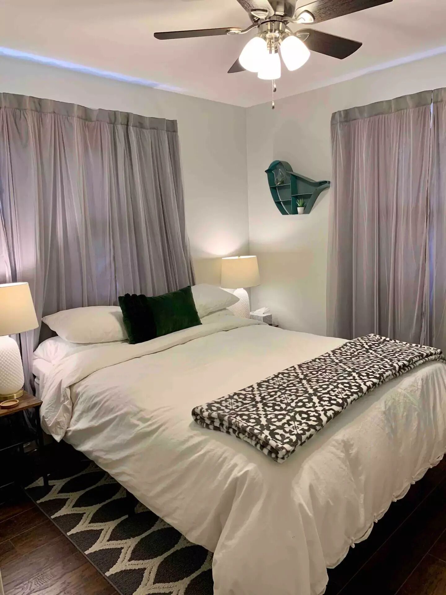 Un lit bien fait, avec du linge de lit blanc et un couvre-lit à motifs, un ventilateur de plafond et une étagère murale agrémentée d’une plante.