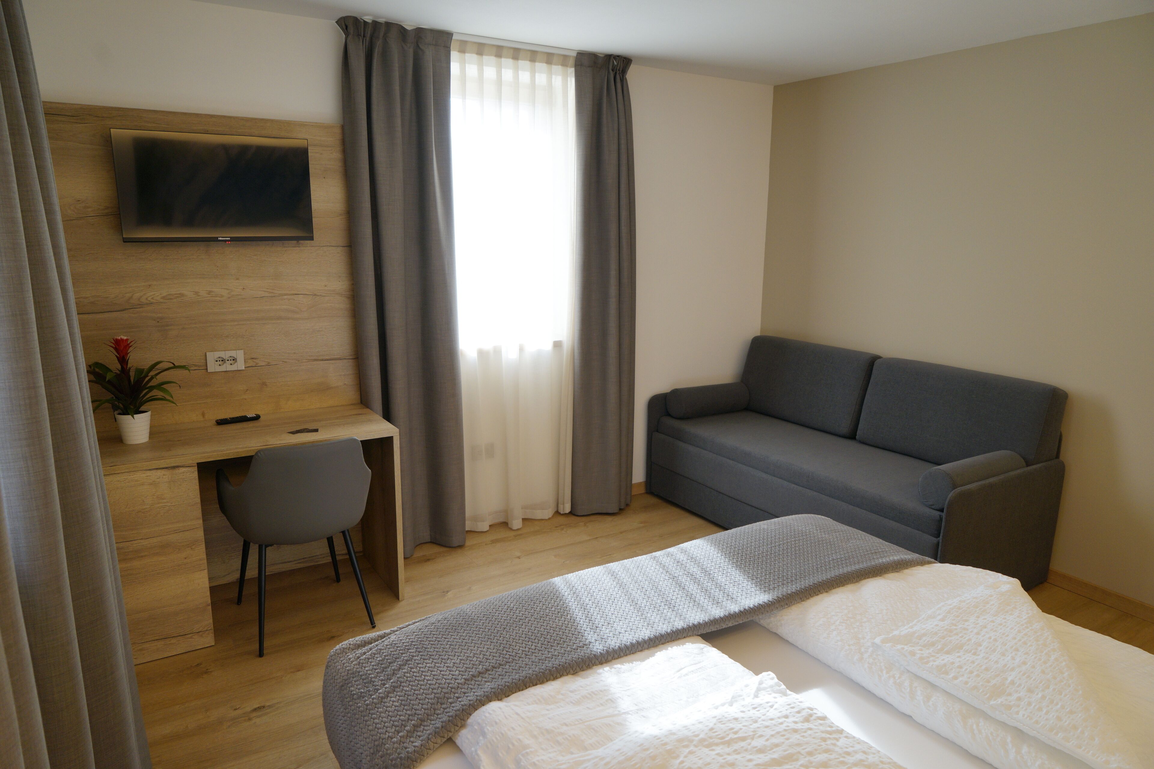 A modern hotel room with a wooden desk, a grey sofa, a bed with a grey blanket, and a TV mounted on the wall.