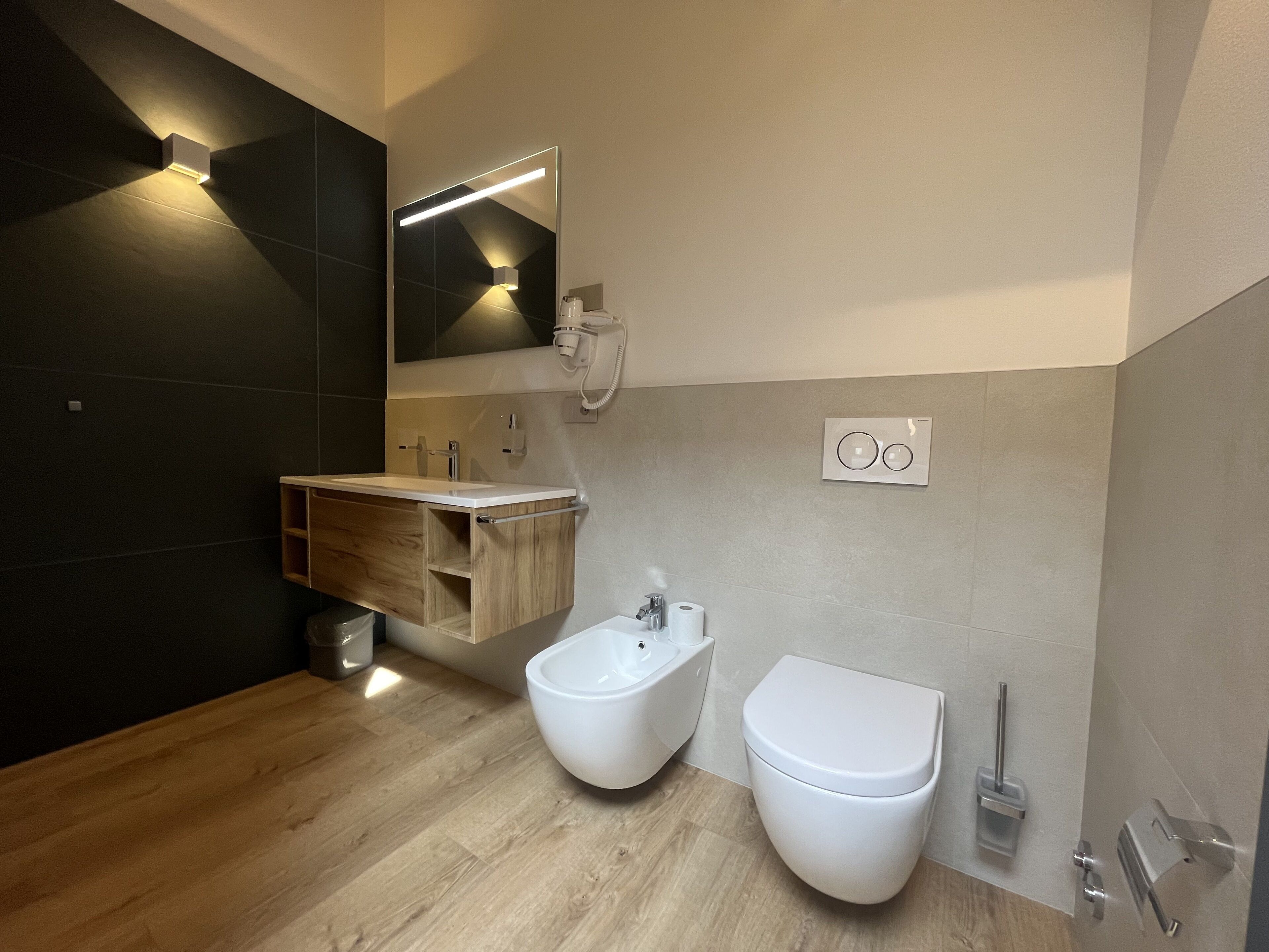 Une salle de bain moderne avec un meuble-lavabo en bois, un bidet, des toilettes et un miroir doté d’un éclairage intégré.
