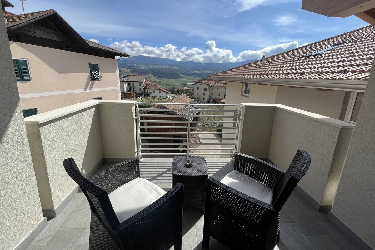 A balcony with two wicker chairs and a small table, overlooking a residential area with houses and a mountain in the background.