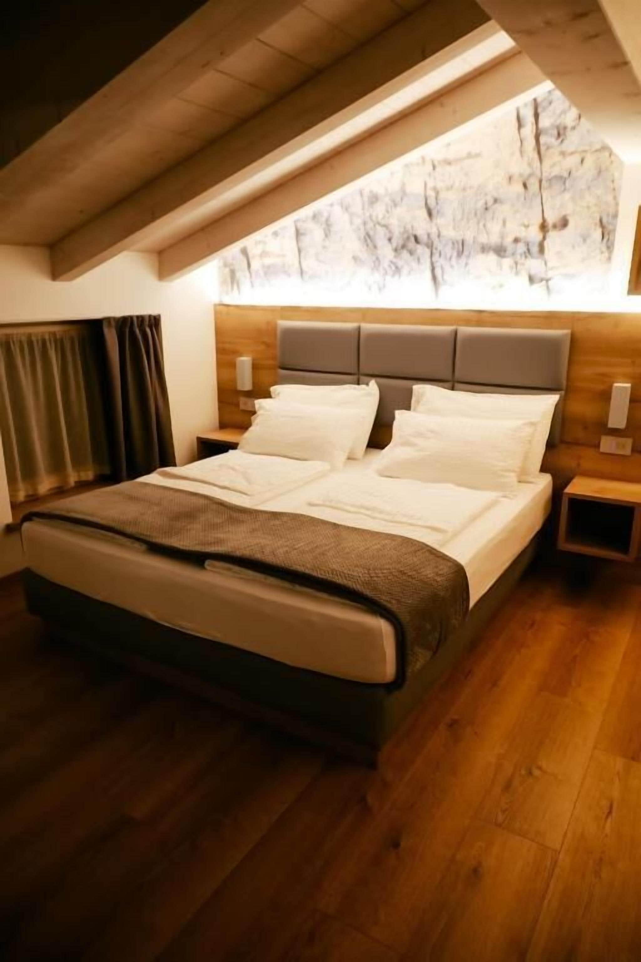 Un dormitorio con techo de madera, una cama con ropa de cama blanca, una mesita de noche y vistas a un paisaje rocoso a través de una ventana.